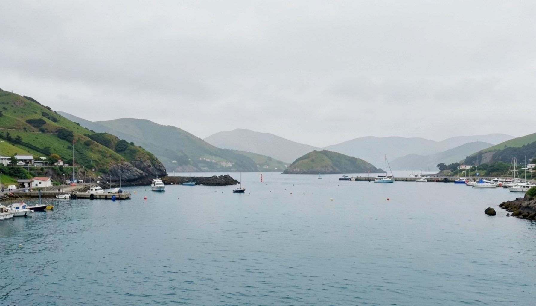 A calm coastal harbour scene with pale grey sky, soft green hills, and muted blue water, giving a quiet island news atmosphere